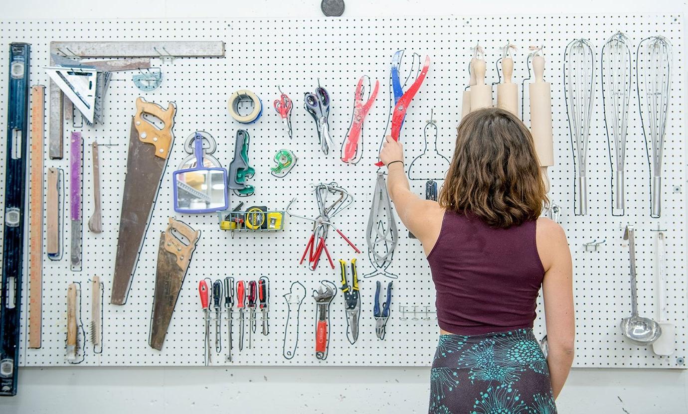 a student chooses a tool hanging from a pegboard in ceramics class. 